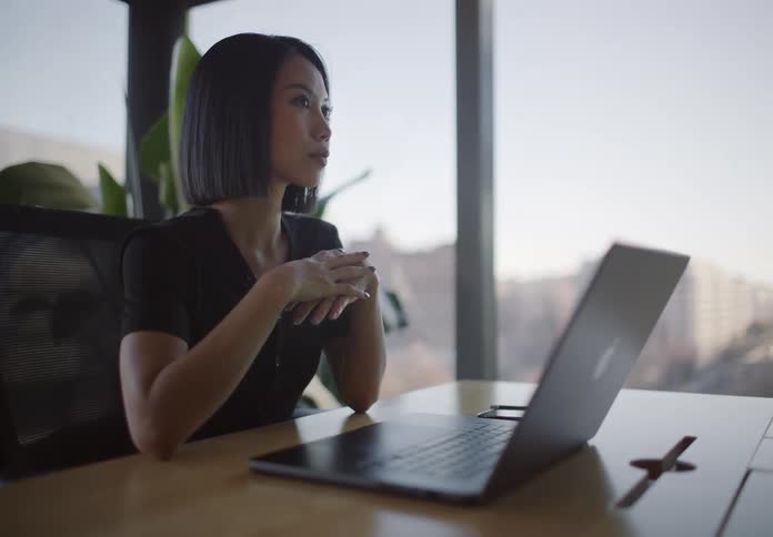 A pensive businesswoman with short hair contemplates while sitting at her desk in a sunlit office overlooking the city