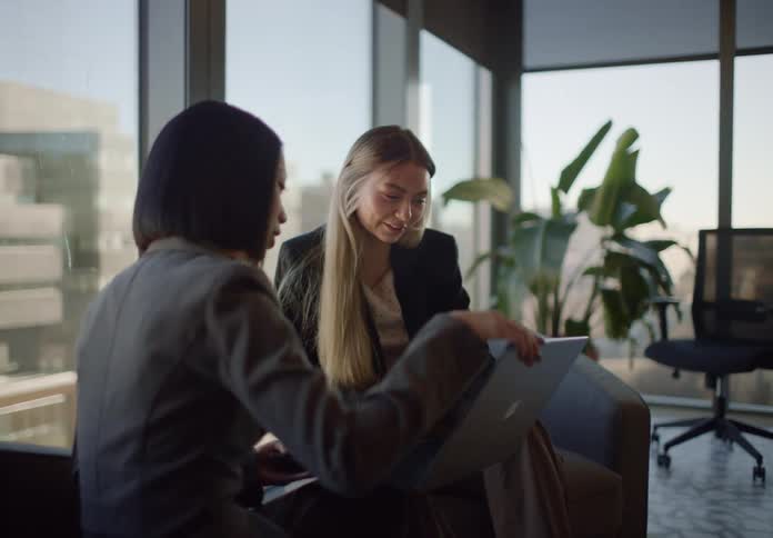 Two diverse businesswomen collaborate on a laptop during a meeting in a modern, sunlit office lounge
