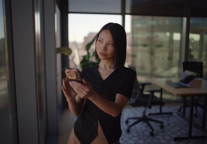 An elegant Asian businesswoman thoughtfully uses her smartphone while standing by a large window in her modern city office