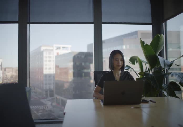 A businesswoman works on her laptop in a modern, sunlit conference room with large windows overlooking the city as the camera slowly pulls back