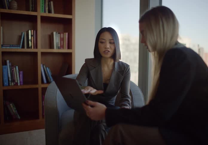 Two diverse businesswomen sit in a modern office lounge and collaborate on a project using a laptop computer