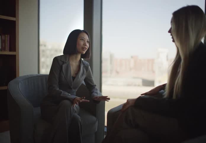An Asian businesswoman gestures as she speaks with a colleague during a meeting in a sunlit office overlooking the city