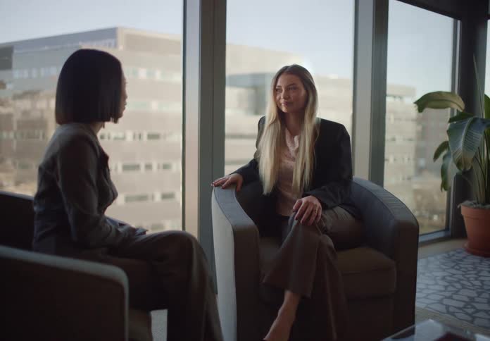 Two diverse businesswomen have an engaging one-on-one conversation while seated in a modern office lounge with large windows