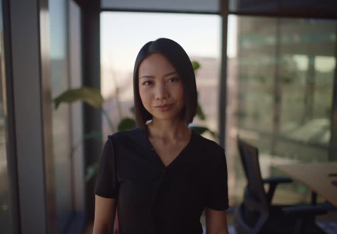A young Asian businesswoman confidently walks towards the camera after sending a message on her phone in a sunlit modern office
