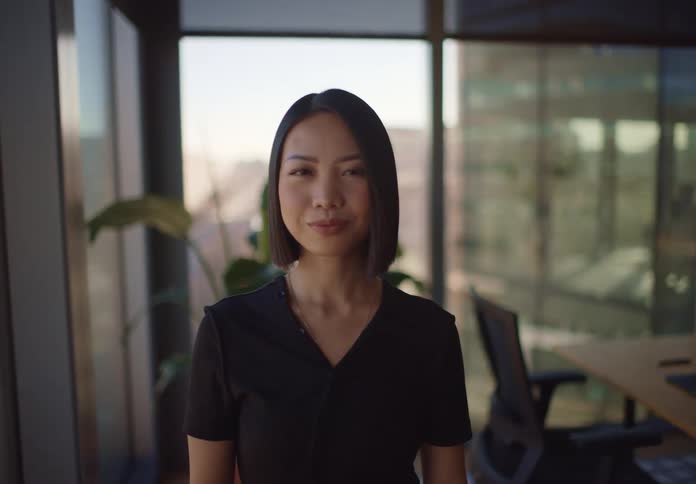 A stylish Asian woman in a modern office looks up from her phone and smiles confidently at the camera