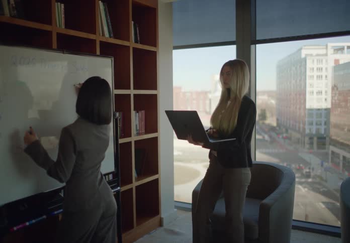 Two businesswomen collaborate on projected sales figures in a modern office with a city view, one writing on a whiteboard and the other holding a laptop