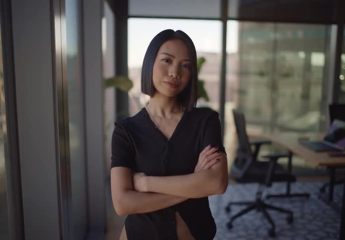 A confident young Asian businesswoman crosses her arms and looks at the camera while standing in a modern, sunlit office