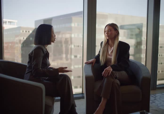 Two diverse businesswomen have a professional discussion in a modern office lounge with a large window showing a city view