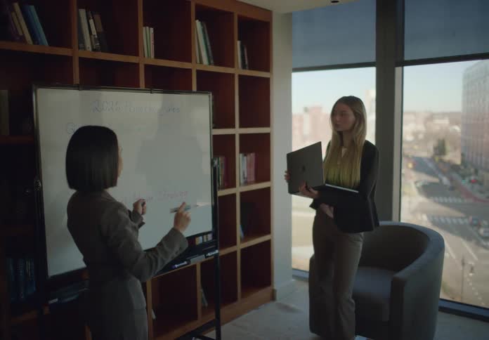 Two businesswomen strategize over projected sales using a whiteboard and a laptop in a modern office with a large window overlooking the city