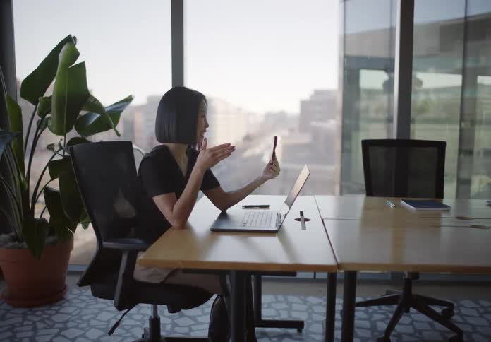 A stylish young woman with a prosthetic leg smiles and waves while on a video call in a modern office with a city view