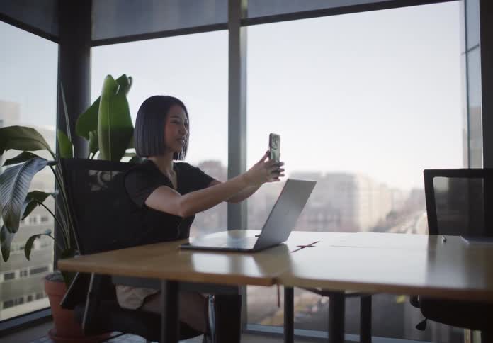 A stylish young Asian woman takes a break from work to video call a loved one from her sunny, modern office, smiling and blowing a kiss