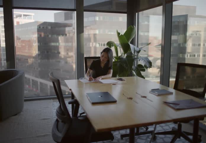 Slow zoom on a focused businesswoman writing at a table in a modern corner office with a city skyline view