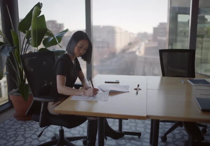Focused young Asian businesswoman signs important documents in a sunlit high-rise office overlooking the city