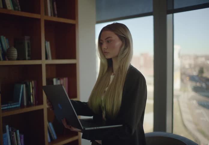 A focused young businesswoman works on her laptop while standing in a modern office library with a city view