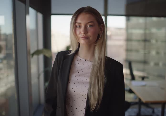 A confident young businesswoman with blonde hair looks up and smiles at the camera in a modern office