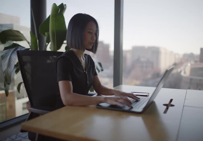 A businesswoman types on her laptop at a desk in a modern office with large windows showing a city view, then checks her phone
