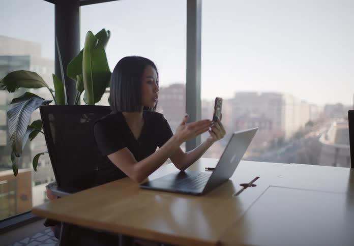 A stylish Asian professional takes a video call on her phone in a sunny, modern office with a city view