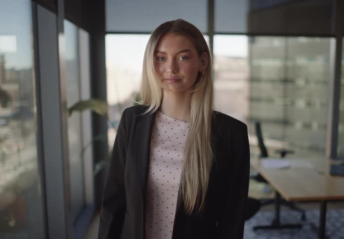 A confident young businesswoman with blonde hair looks up and smiles at the camera while standing in a modern office