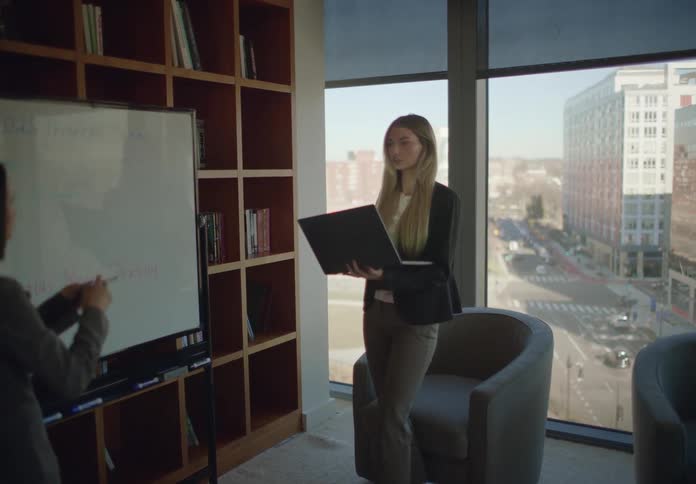 Two businesswomen collaborate on a strategy presentation in a modern, sunlit office with a city view