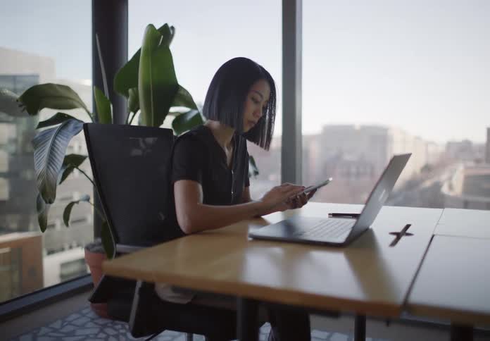 A focused Asian businesswoman analyzes data on her smartphone and laptop in a modern high-rise office overlooking the city