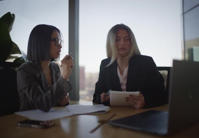 Two diverse female colleagues discuss business strategy in a modern office, reviewing information on a laptop
