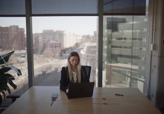 A professional woman works on her laptop at a desk in a modern office with a view of the city as the camera slowly zooms in