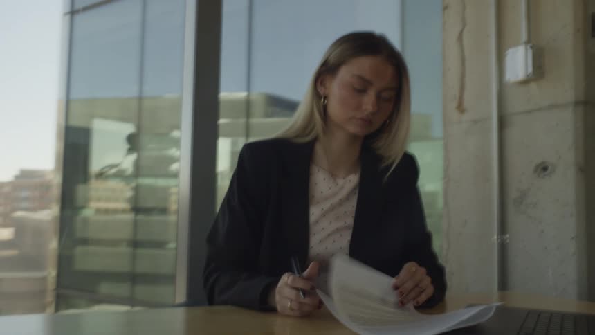 A young businesswoman sits at her desk in a modern office, diligently reviewing and signing paperwork with a cityscape visible through the window