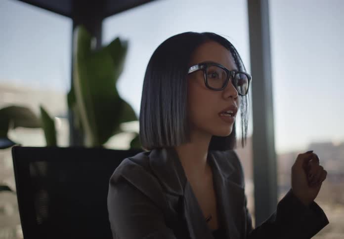 A focused businesswoman in glasses passionately discusses ideas and gestures with a pen in a modern office with a city view