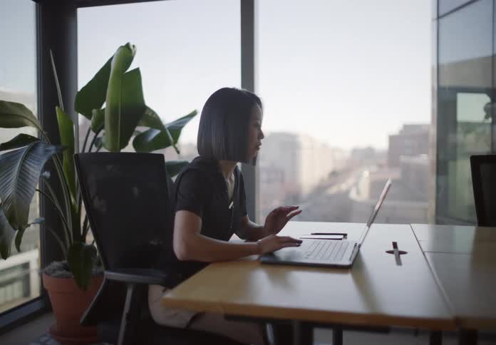 An Asian businesswoman smiles while working on her laptop in a modern corner office with a bright city view