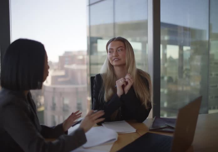 Two professional women collaborate on a project, reviewing documents and using a laptop in a modern high-rise office with a city view