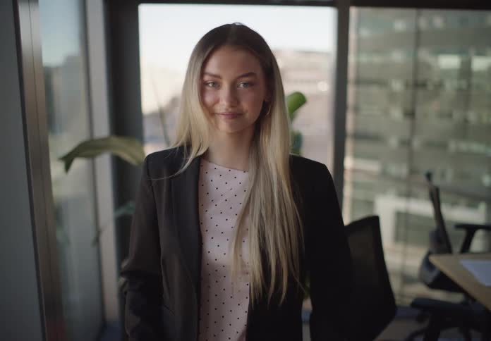 A young, confident businesswoman in a modern city office uses her smartphone before walking forward and smiling at the camera