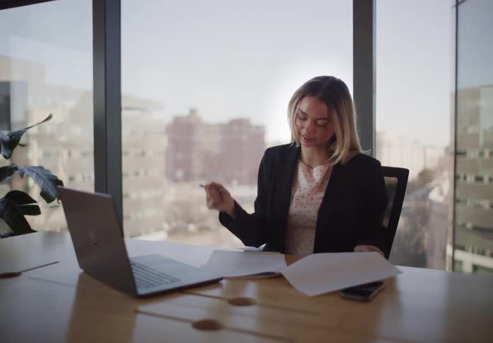 A focused businesswoman writes notes and reviews paperwork at her desk in a modern office with a city view