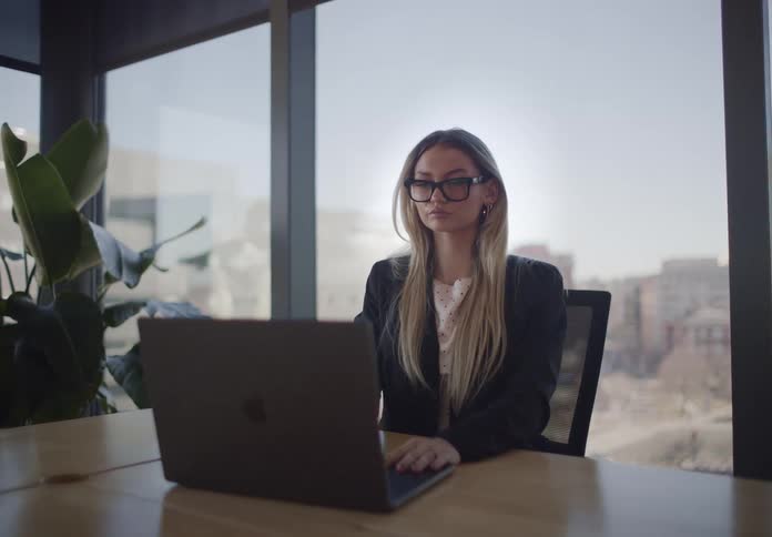 A focused businesswoman works on her laptop in a modern office with a panoramic city view