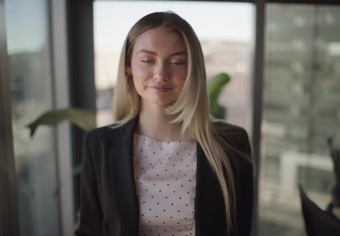A confident young businesswoman walks toward the camera and smiles in a bright, modern office space