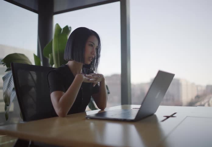 A thoughtful Asian businesswoman works on her laptop in a modern corner office, pausing to contemplate while looking out at the city skyline