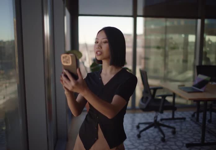 A stylish Asian businesswoman smiles while on a video call on her smartphone, standing by a large window in a modern, sunlit office