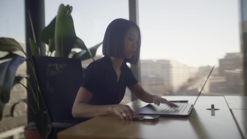 A focused Asian businesswoman works on her laptop and checks her phone in a modern office with a city view