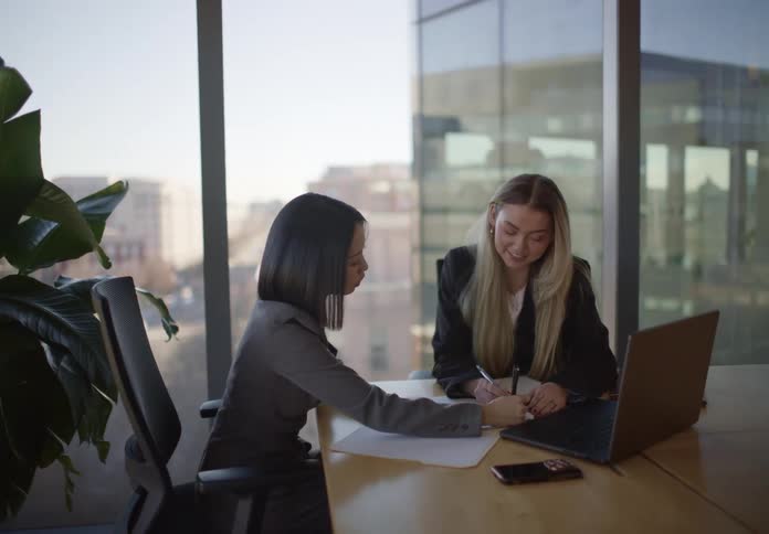 Two professional businesswomen collaborate, discussing and signing a contract together in a modern city office with a large window view