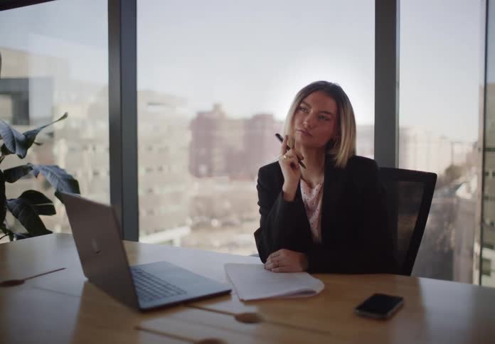 A thoughtful businesswoman writing notes at her desk in a modern city office pauses to think, then smiles as she gets an idea and continues writing