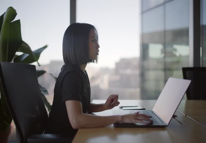 A focused Asian woman works on her laptop in a modern, sunlit office with a panoramic city view