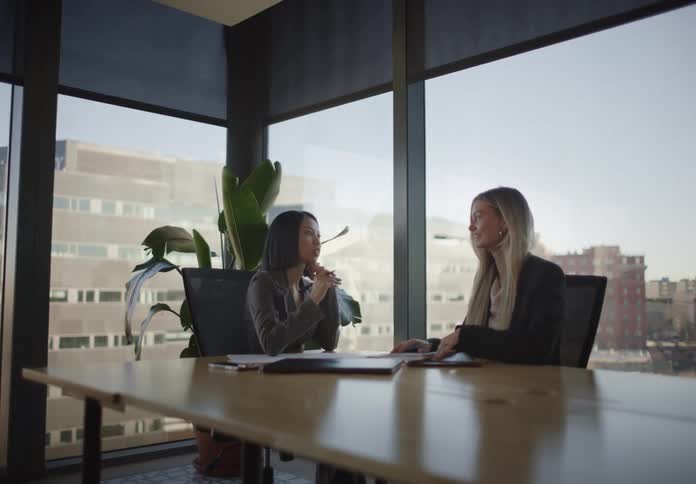 A slow push-in on two professional women having a business discussion in a modern office conference room with large windows overlooking the city