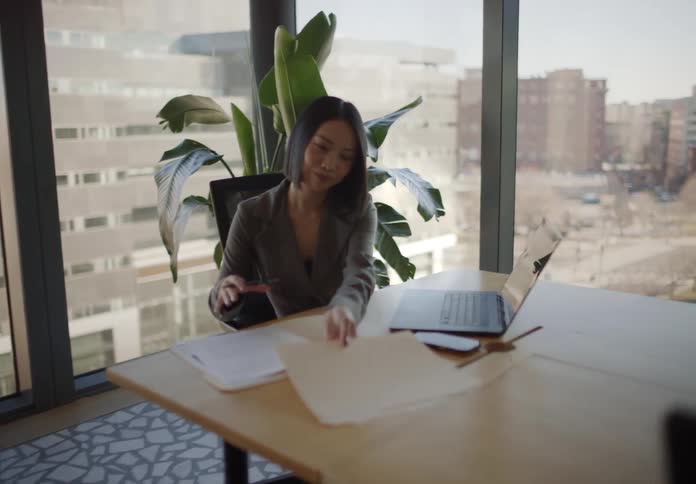 A businesswoman drops off a folder and does a happy dance while her colleague works at her desk in a sunlit high-rise office