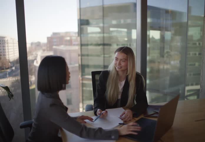 Two professional women collaborate during a business meeting in a modern office with large windows overlooking the city