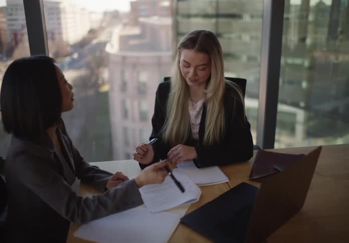 Two professional businesswomen collaborate in a modern city office, discussing and reviewing documents together