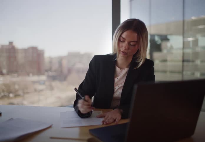 A blonde businesswoman in a black blazer sits at her desk by a large window and diligently writes on paperwork