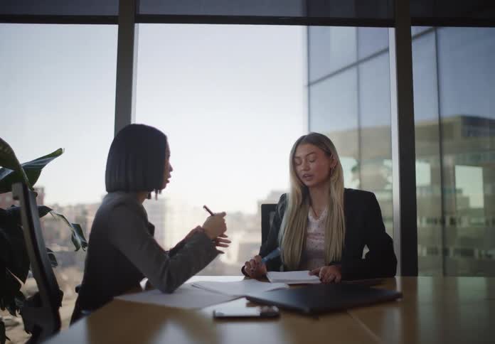 Two diverse businesswomen have a focused discussion in a modern conference room with a large window overlooking the city