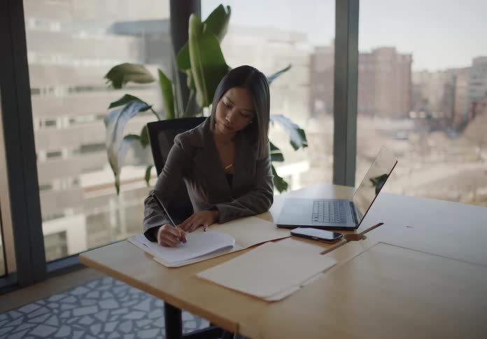 A business professional receives a folder from her colleague and begins working at her desk in a modern, sunlit office with a city view