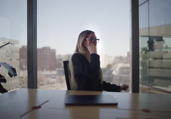 A young businesswoman finishes her work, closes her laptop, and takes a moment to thoughtfully look out her office window at the city