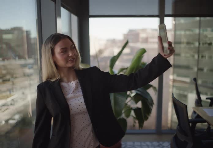 A young businesswoman films a selfie video or takes a video call in a modern office overlooking the city