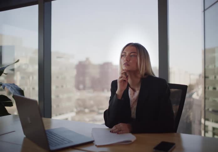 A pensive young businesswoman pauses her work to reflect and gaze out the window in a modern, sunlit city office